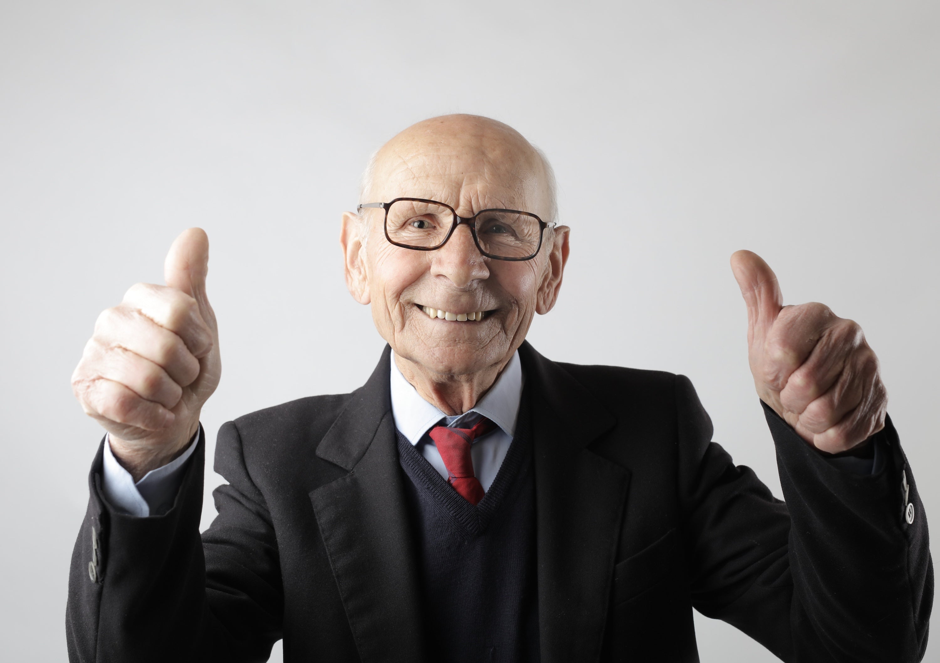 Older man in a suit and tie wearing glasses giving a thumbs-up gesture against a plain background